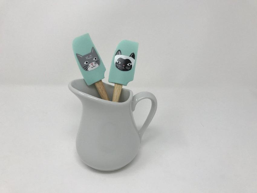 Pair of wooden handle spatulas with turquoise green heads featuring cats on them inside of a white gravy boat 