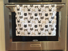 Load image into Gallery viewer, White cat-themed kitchen dish towel featuring numerous cats on it hanging from an oven handle bar
