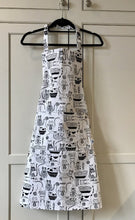 Load image into Gallery viewer, Picture of a white and black kitchen apron on a black hanger featuring black cats 
