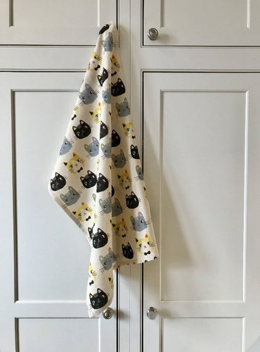 Off-white colored cat-themed dish towel featuring numerous cats on it hanging from a white kitchen cupboard handle