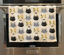 Load image into Gallery viewer, Off-white cat-themed kitchen towel featuring numerous cats on it hanging from an oven handle bar
